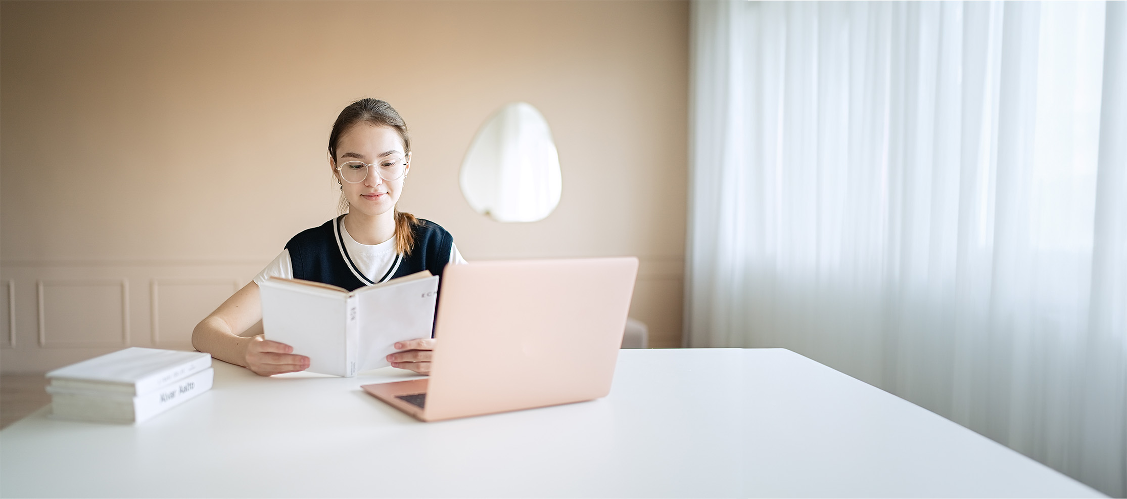 Girl reading book with laptop in front of her